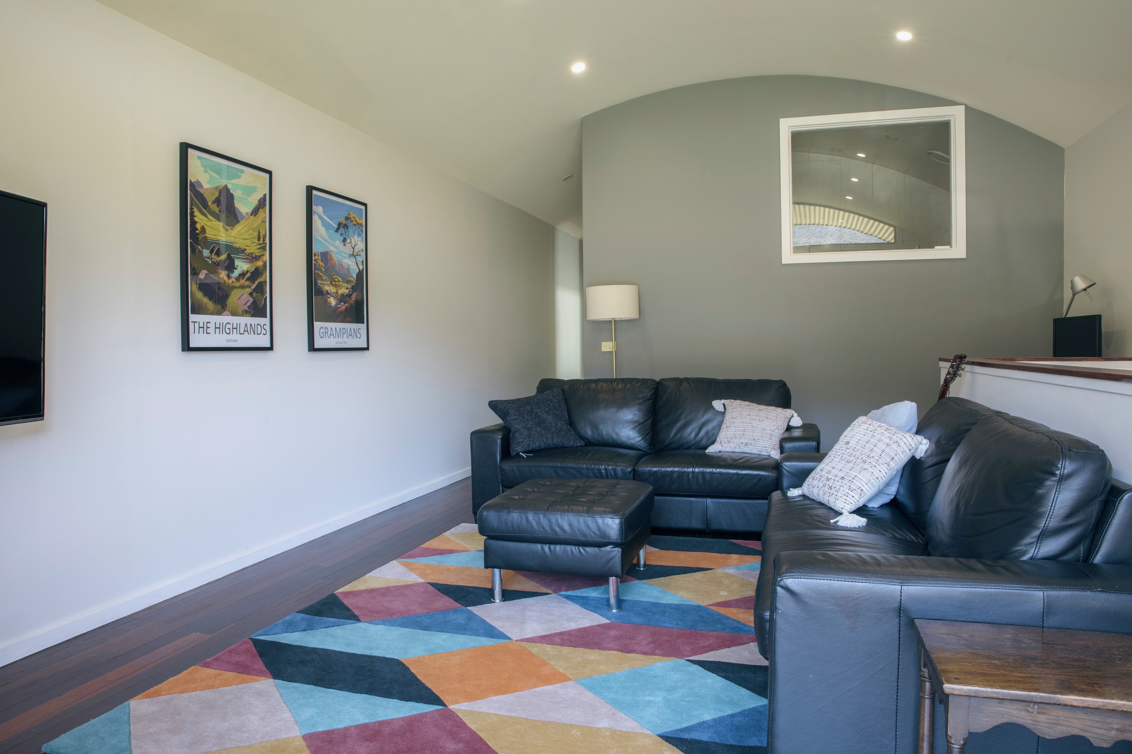 A modern living room with a black leather sofa, a colorful geometric rug, and two framed pictures on the wall.