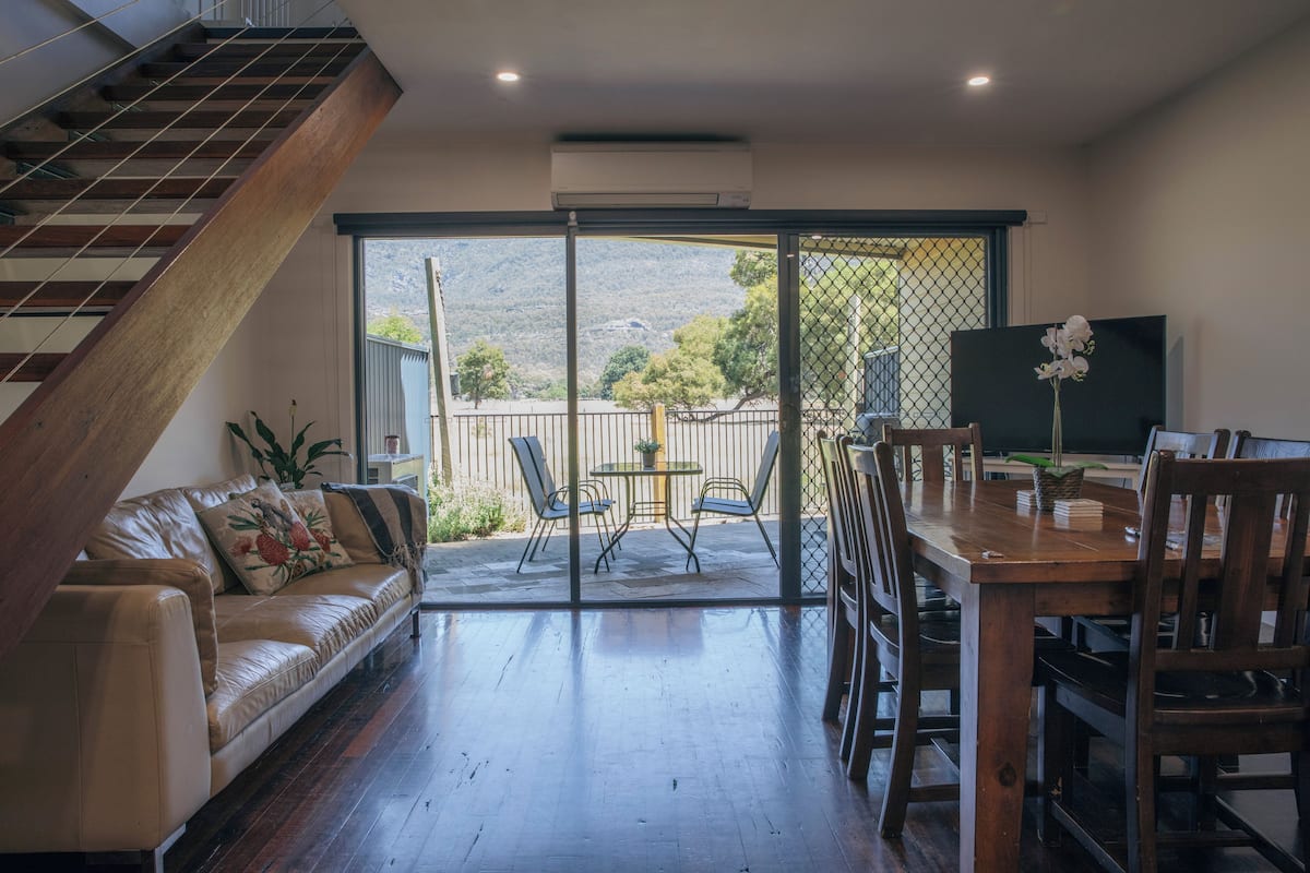A spacious living area with a wooden staircase, a leather sofa, a dining table with chairs, and a large sliding glass door leading to an outdoor patio.