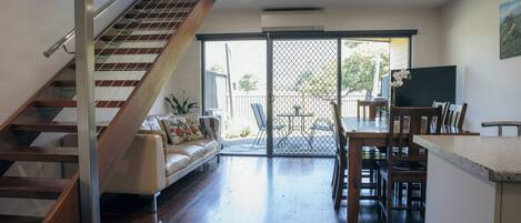 Un intérieur moderne avec un escalier en bois, un canapé, un coin repas et une porte-fenêtre coulissante donnant sur un espace extérieur.