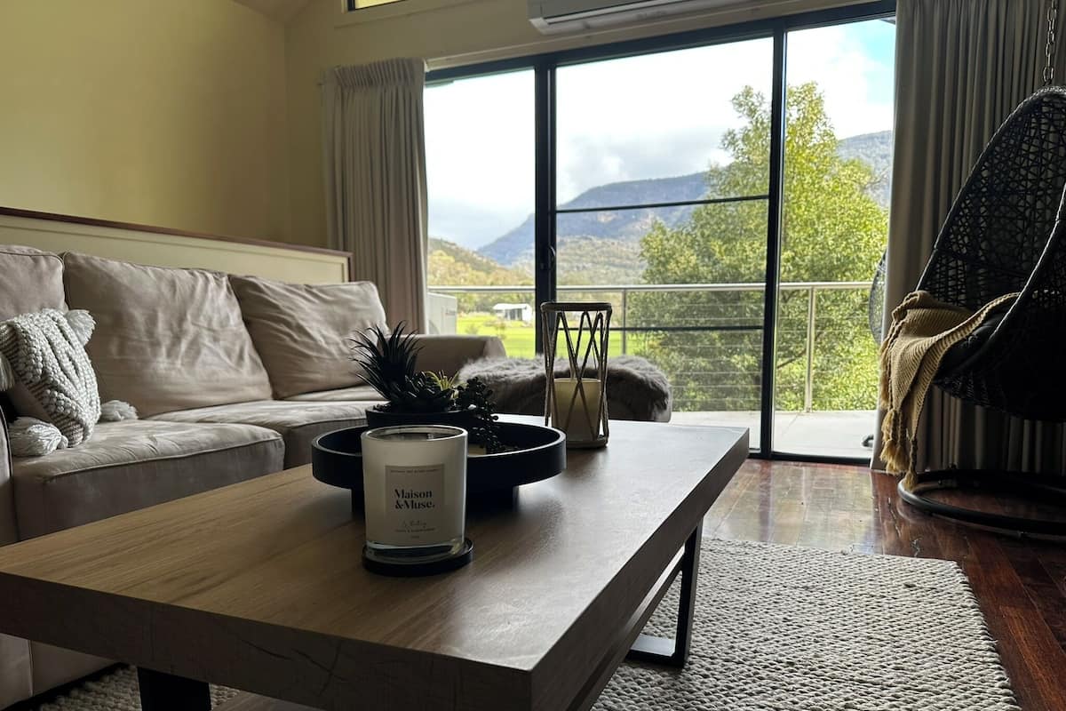 A modern living room with a sofa, a coffee table with a candle, and a view of a mountainous landscape through large windows.
