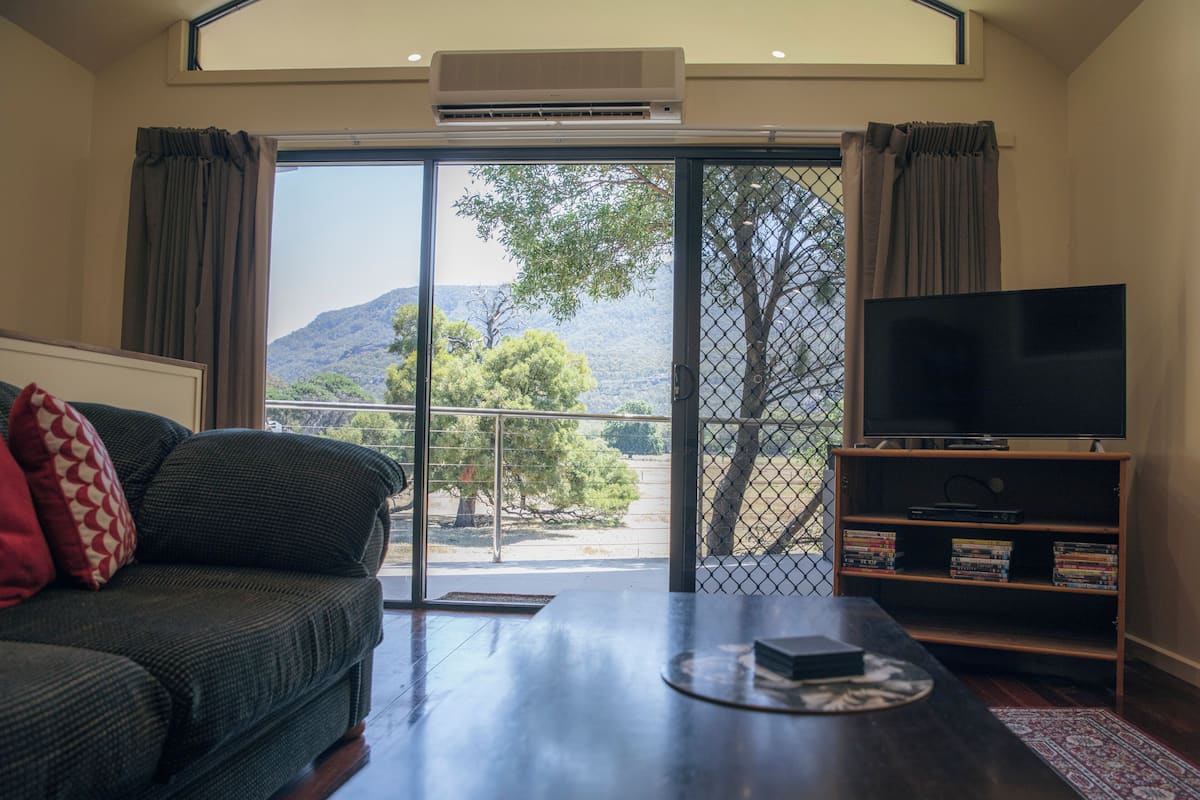A living room with a sofa, a coffee table, a TV, and a bookshelf. There is a large window with a view of trees and hills.