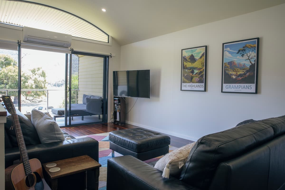 A living room with a black leather sofa, a wooden coffee table, and a guitar. There is a flat-screen TV mounted on the wall and two framed posters of landscapes.