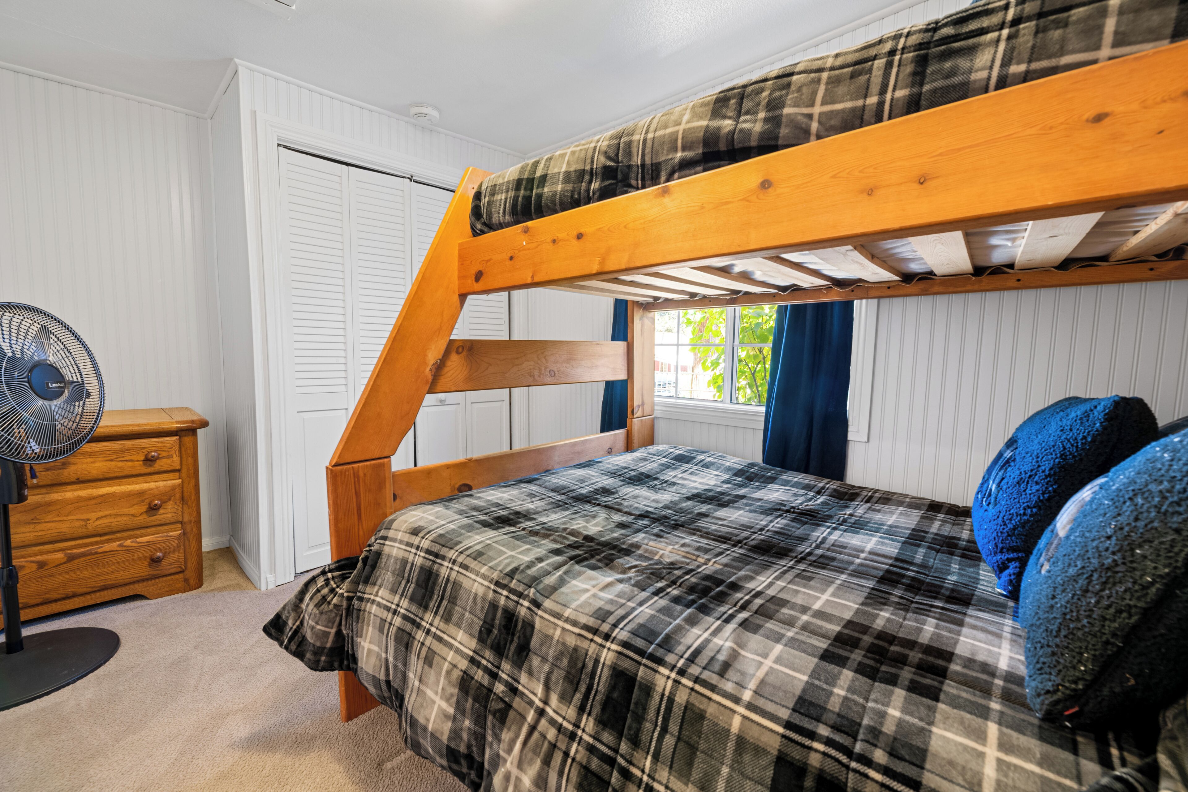Una cama individual superpuesta con colcha escocesa, un aparador de madera y una ventana con cortinas.