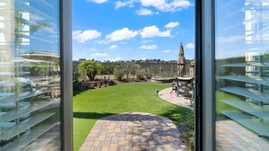 A view from a modern building with large glass windows, a well-maintained lawn, and a fenced garden area. - Spacious w/ Yard, BBQ, Presidio Access Near Park, Pool, Tennis, Beaches & Legoland! Extended Stays (Carlsbad)