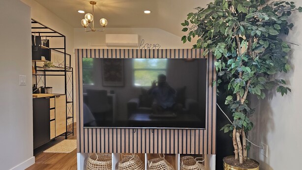 Sala de estar con un televisor de pantalla plana grande montado en una pared a rayas, una planta en maceta y un aparador con objetos decorativos.