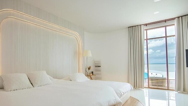 A modern hotel room with a large bed, a bedside table with a lamp, and a view of the ocean through large windows. - BanyakasSirindhron (Sirindhorn)