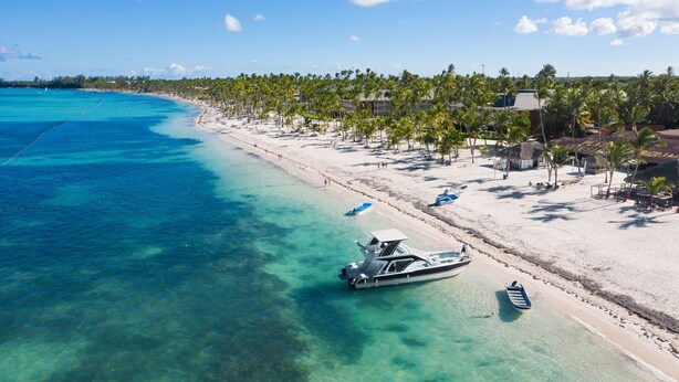 Une plage aux eaux turquoise et limpides, un bateau amarré sur le rivage et une rangée de palmiers bordant la plage.