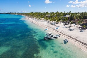A beach with clear turquoise waters, a boat docked on the shore, and a row of palm trees along the beach.