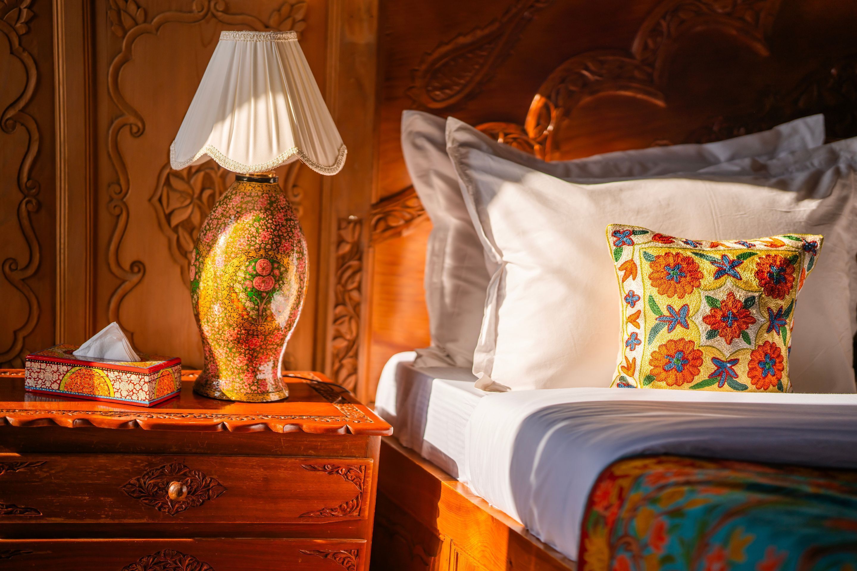 A bed with white linens, a colorful floral pillow, a decorative lamp, and a wooden nightstand with a tissue box.