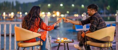 Duas pessoas sentadas à mesa com uma vela, com vista para um corpo de água e luzes ao fundo.