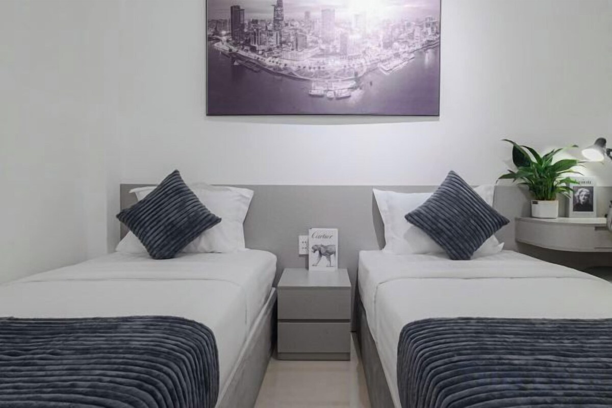 Een hotelkamer met twee bedden, een nachtkastje, een bijzettafeltje met een lamp, een ingelijste foto aan de muur en een potplant.