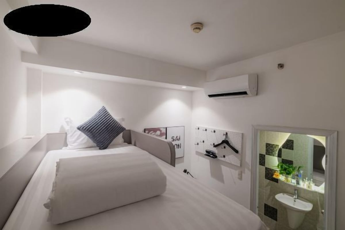 A modern hotel room with a large bed, a bedside table, a wall-mounted air conditioning unit, a small bathroom with a sink and mirror, and a wall with a grid pattern and a hanging coat rack.