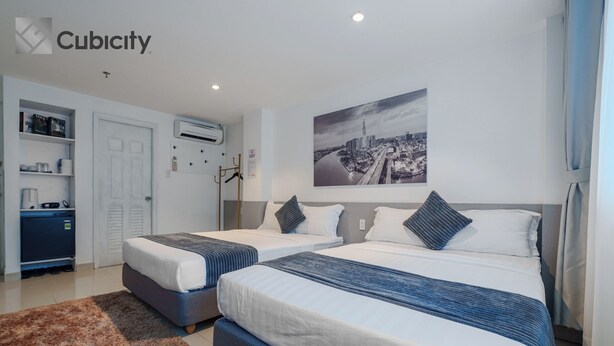 A modern hotel room with two beds, a cityscape artwork, a wardrobe, and a desk with a computer. - Cubicity De Tham (Ho Chi Minh City)