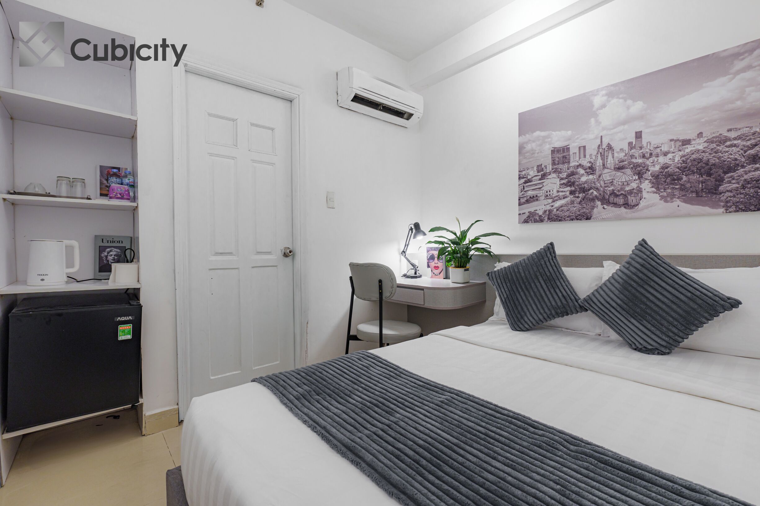 A hotel room with a bed, a desk, a chair, a small refrigerator, and a wall-mounted air conditioner.