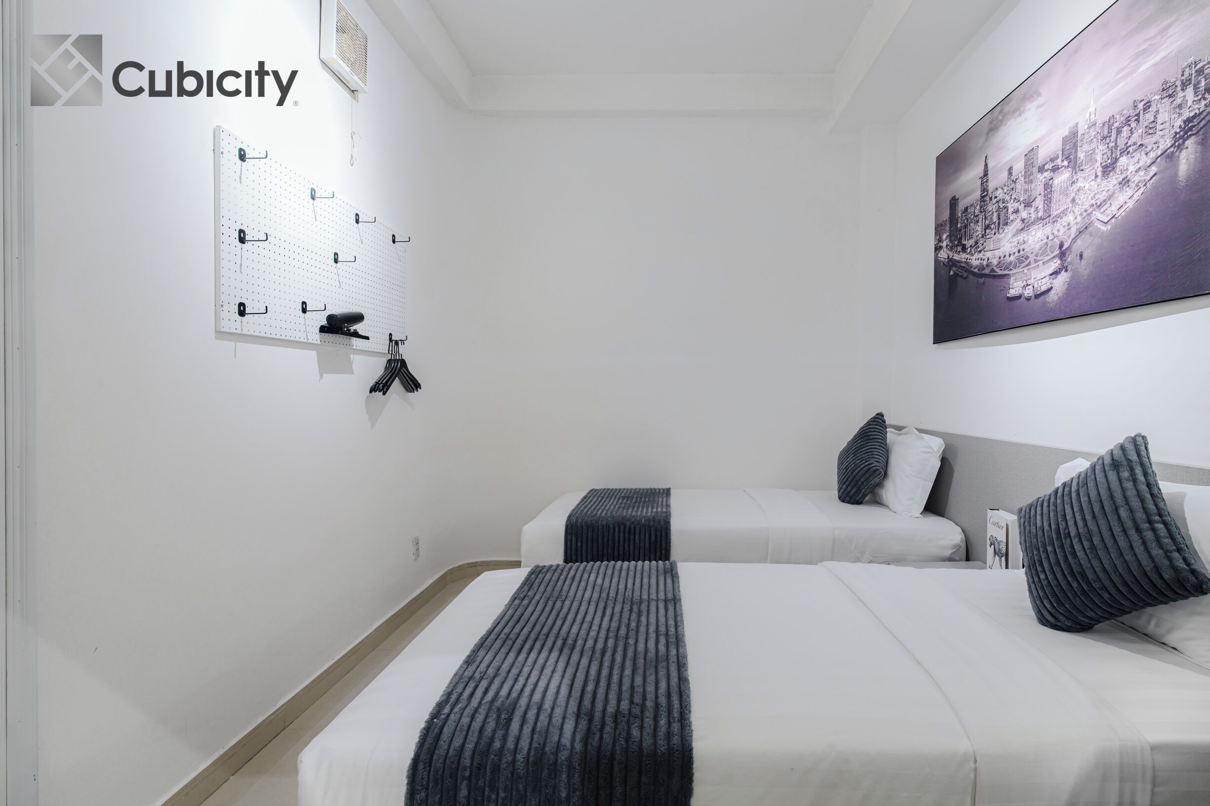 Une chambre d’hôtel avec deux lits, un mur recouvert d’un panneau perforé et un tableau représentant un paysage urbain.