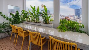 A balcony with a long white table and yellow chairs, surrounded by potted plants and overlooking a cityscape. - Cubicity De Tham (Ho Chi Minh City)