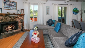 A living room with a stone fireplace, a grey sofa with blue cushions, a wooden coffee table, and a view of the outdoors through large windows. - Bayview Condo (Big Bear Lake)