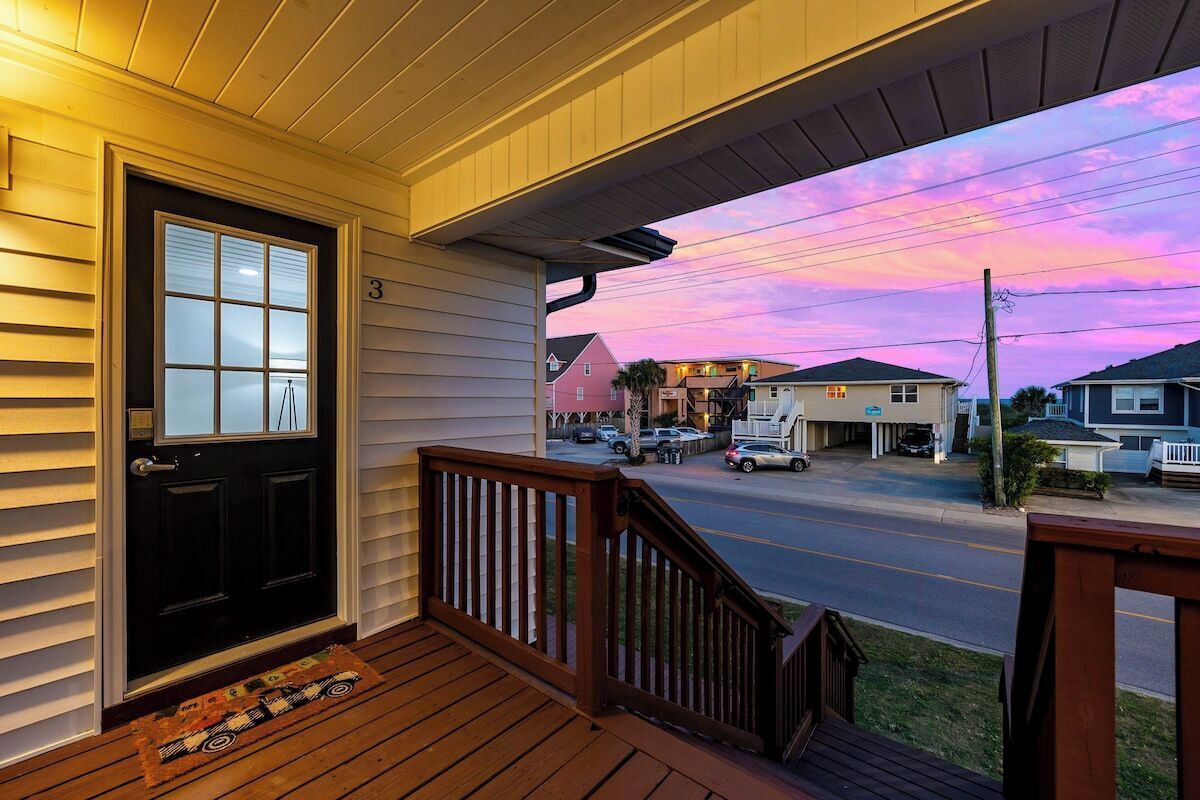 A porch with a black door, a wooden railing, and a view of a street with houses and parked cars.