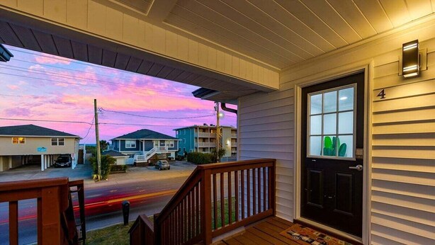 A porch with a wooden railing, a black door with a number, and a view of a street with houses. - Bright 2BR Surf Hideaway | 1-Min Walk to Waves (North Myrtle Beach)