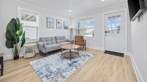 A living room with a grey sofa, a round coffee table, a potted plant, and a TV mounted on the wall. - 2BR Modern Surf Retreat | Steps to Sand (North Myrtle Beach)