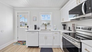 A modern kitchen with white cabinetry, a dishwasher, an oven, a microwave, and a window with blinds. - 2BR Modern Surf Retreat | Steps to Sand (North Myrtle Beach)