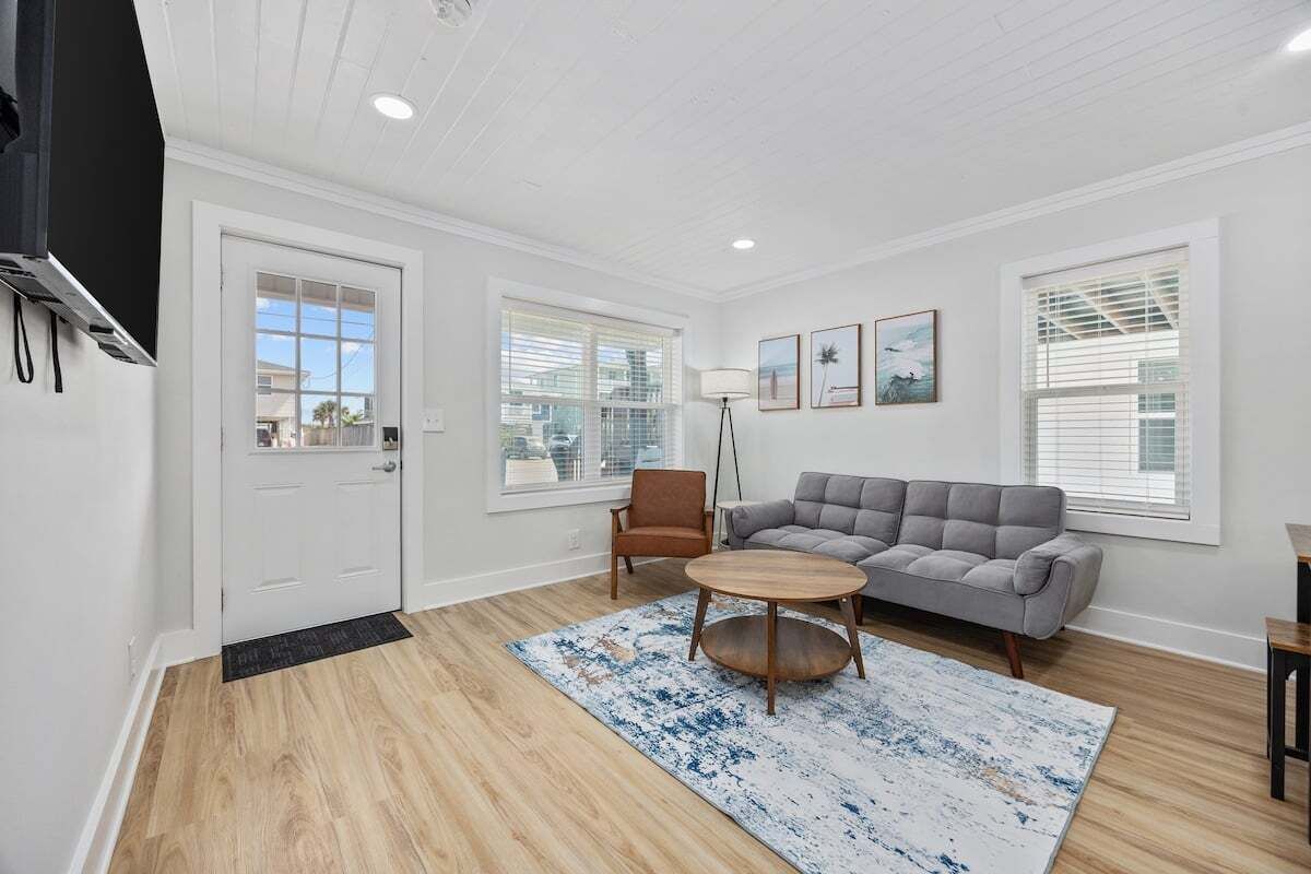 A living room with a gray sofa, a round coffee table, and a brown armchair. There are two windows with blinds, a door, and a lamp.