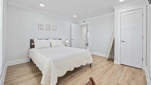 Un dormitorio con cama, paredes blancas, suelo de madera, una puerta y una mesita con lámpara.