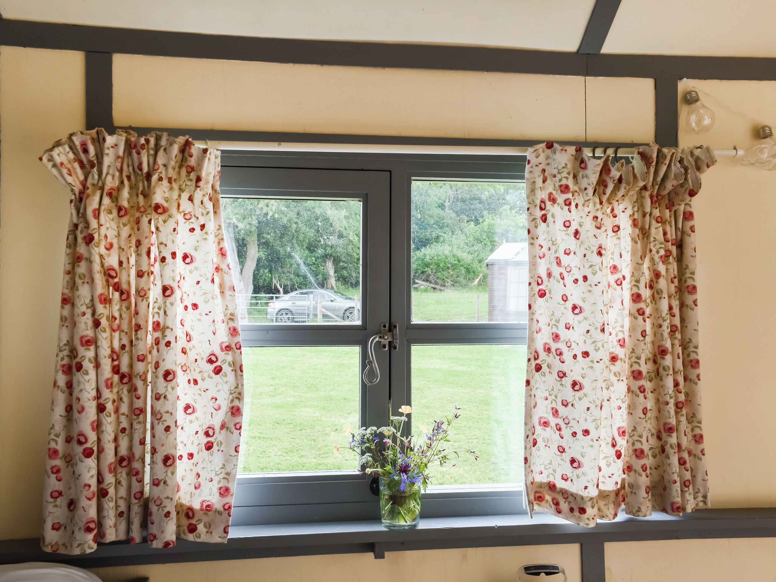 Ein Fenster mit floralem Vorhang, ein kleiner Blumenstrauß in einer Vase und Blick auf eine grüne Rasenfläche sowie ein Gebäude im Freien.