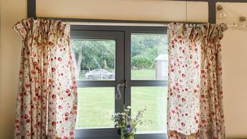 Ein Fenster mit floralem Vorhang, ein kleiner Blumenstrauß in einer Vase und Blick auf eine grüne Rasenfläche sowie ein Gebäude im Freien.