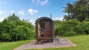 A small wooden structure with a door, a window, and a chimney, surrounded by a gravel area and grass, with trees in the background. - Brown Hare - UK49799 (Brecon)