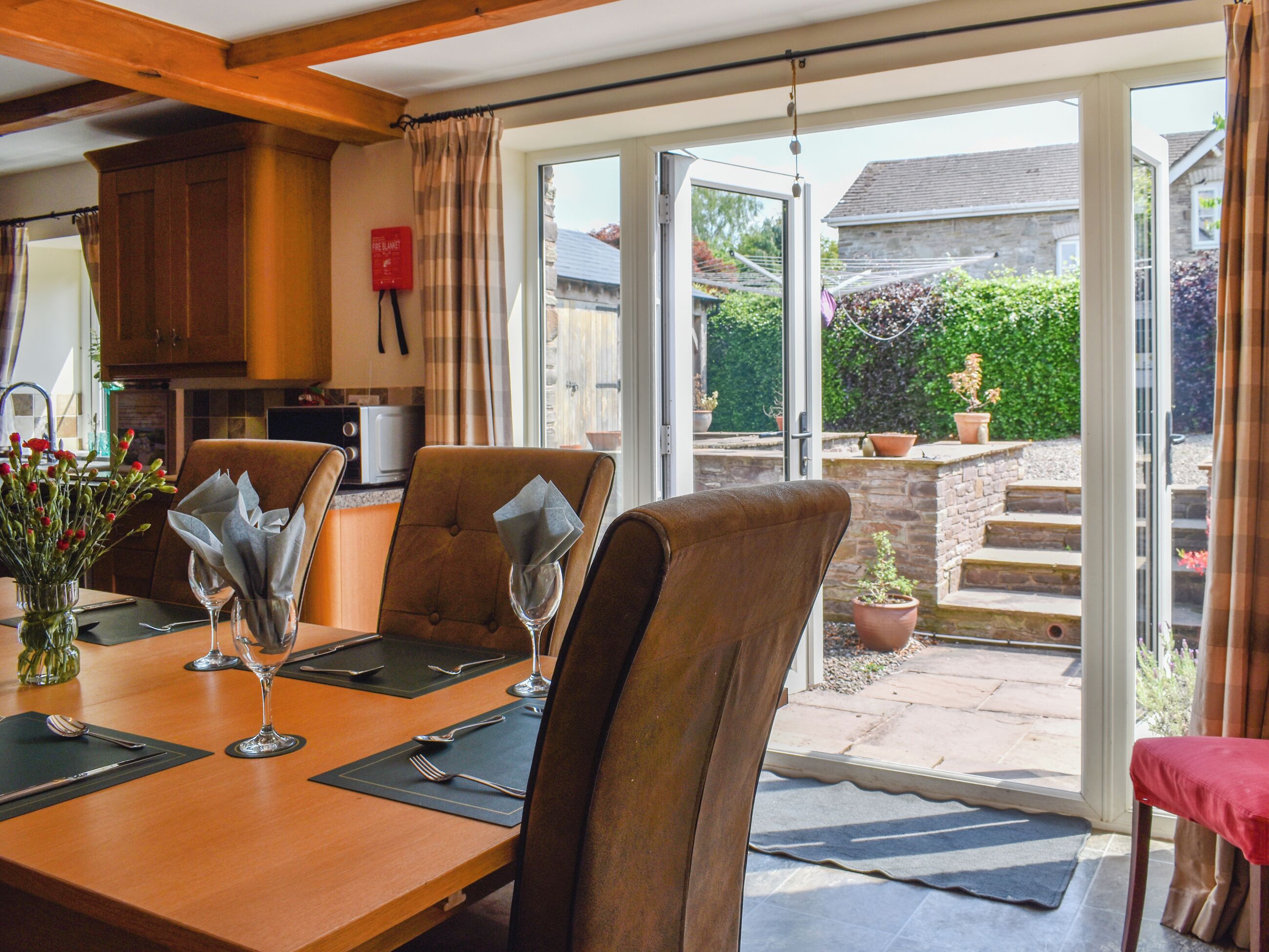 A dining area with a table set for four, chairs, a vase with flowers, and a view of the garden through glass doors.