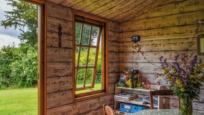 Interior rústico de una cabaña de madera con un ventanal grande, una mesa con mantel floral y un estante con juegos de mesa.