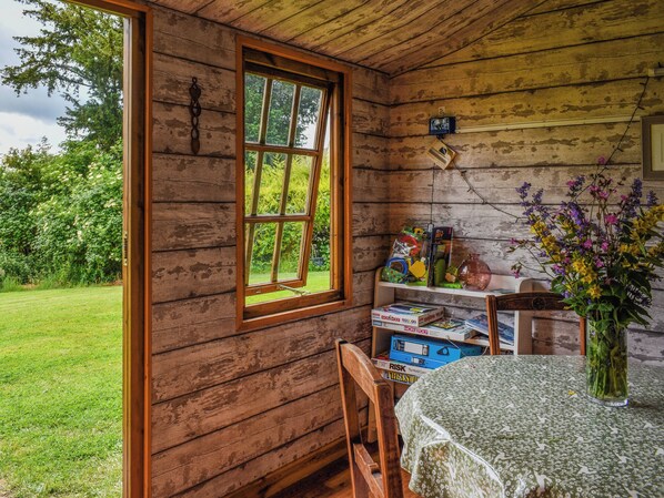 A rustic wooden cabin interior with a large window, a table with a floral tablecloth, and a shelf with board games.