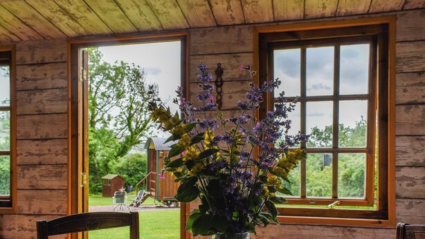 Una cabaña rústica de madera con una mesa redonda y sillas, un jarrón de flores y vistas a un paisaje verde a través de ventanas amplias.