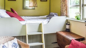 Una habitación pequeña y acogedora con una cama litera, una ventana con cortinas, un sillón de mimbre y una mesita.