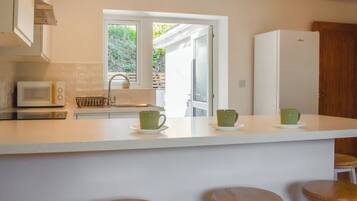A kitchen with a white countertop, a microwave, a sink, and a window. There are three stools and a refrigerator.