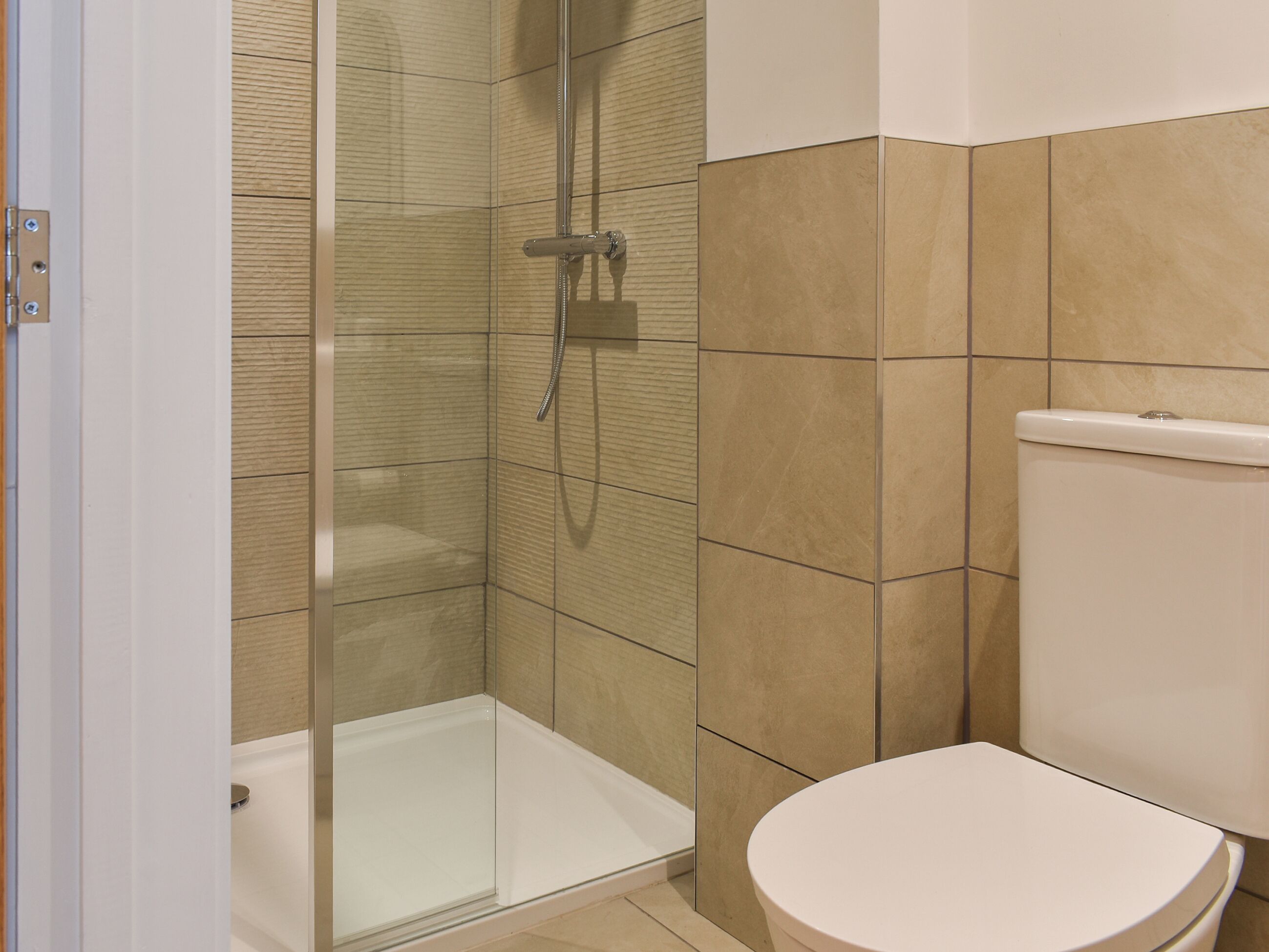 Un baño con ducha envuelta en vidrio, inodoro blanco y paredes con azulejos color beige.