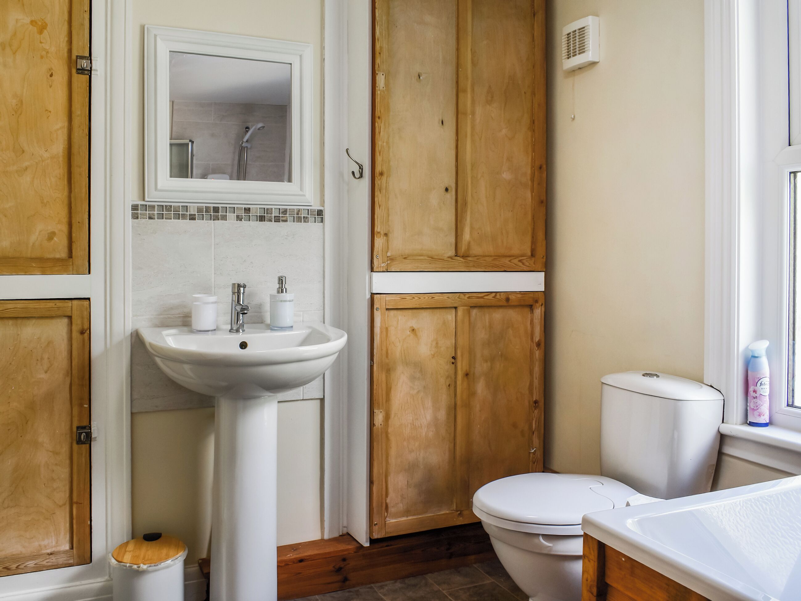Een badkamer met een wit wastafel, een spiegel, houten kastjes, een toilet en een raam.