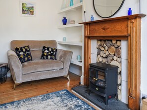Ein Wohnzimmer mit einem beigen Sofa, zwei gemusterten Kissen, einem blau-weißen Teppich, einem schwarzen Kaminofen und einem Kamin mit Holzstapel.