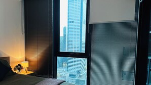 A hotel room with a large window, a bed, a nightstand with a lamp, and a view of tall buildings. - 2 Bedroom Apartment near Southern Cross Station (Melbourne)