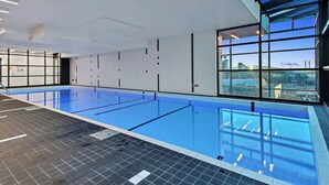 An indoor swimming pool with clear blue water, a tiled floor, and large windows offering a city view. - 2 Bedroom Apartment near Southern Cross Station (Melbourne)