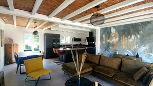 Un soggiorno moderno con soffitto in legno, un divano grande, una zona pranzo e una cucina con vista sul verde.