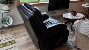 Una sala de estar moderna con un sillón de cuero, un televisor de pantalla plana y un ventanal que ofrece vistas a un paisaje verde.