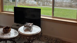 Una habitación con un ventanal grande, un televisor de pantalla plana, dos mesitas redondas y una alfombra mullida.
