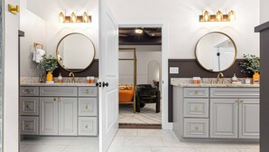 A bathroom with two sinks, each with a round mirror and wall-mounted lighting. The room has a tiled floor and a door leading to another room.