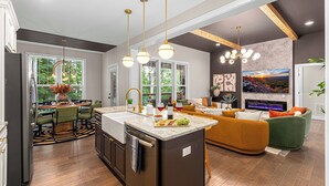 A modern kitchen with a central island, dining area with chairs, and a living room with a sofa and fireplace.