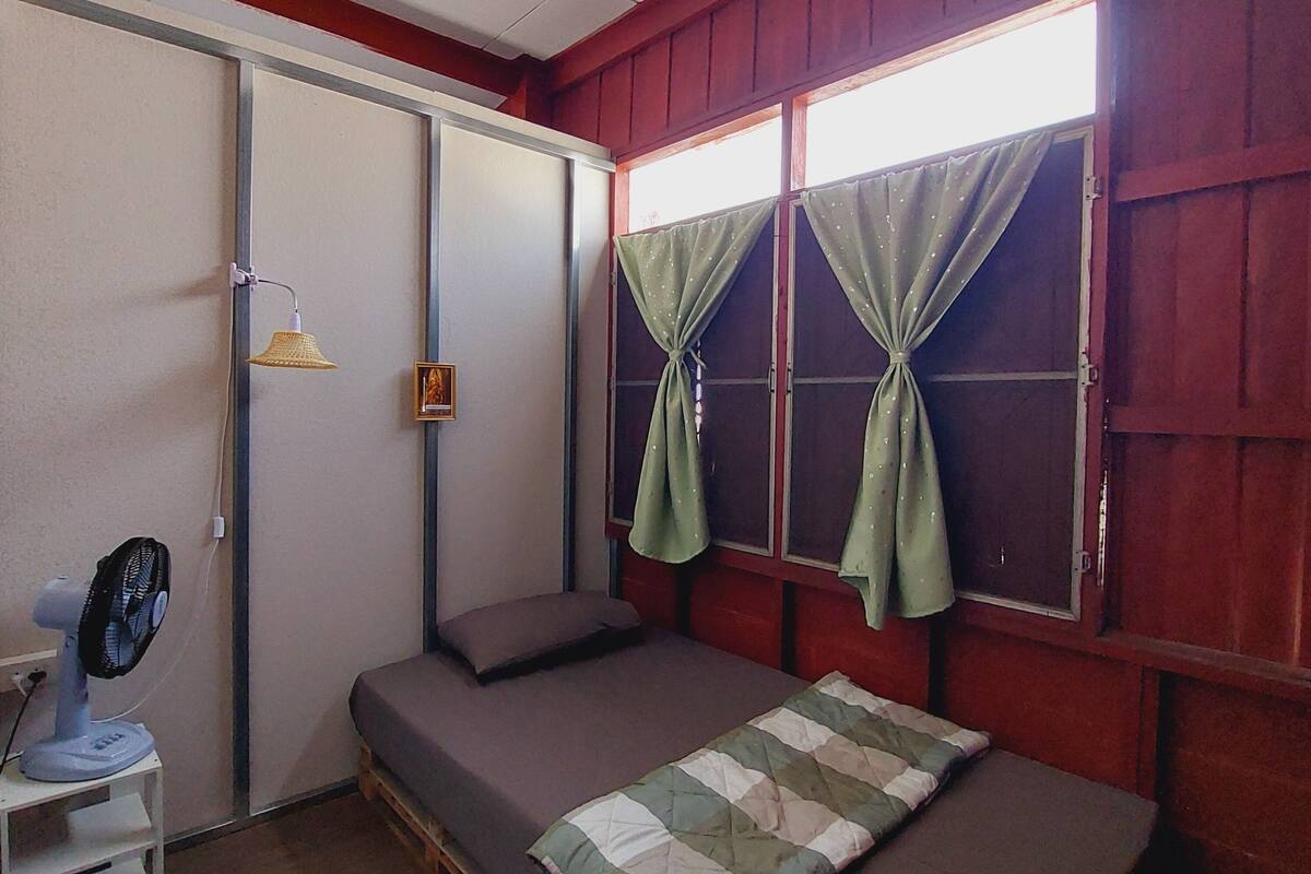 Une chambre de taille modeste, avec un lit simple, un petit bureau équipé d’un ventilateur et une fenêtre avec des rideaux.