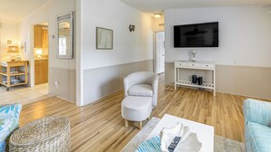 Un salón con un televisor de pantalla plana, un sillón gris, un reposapiés de mimbre y una mesa de centro con un control remoto.
