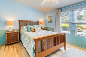 A bedroom with a wooden bed, two bedside tables with lamps, a window with blinds, and a view of greenery outside. - 2 Blocks to Crystal Coast: Family-Friendly Retreat (Harkers Island)