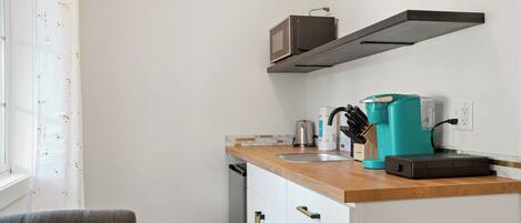 Cocina compacta con armarios blancos, encimera de madera, microondas, cafetera y ventana con cortina blanca.